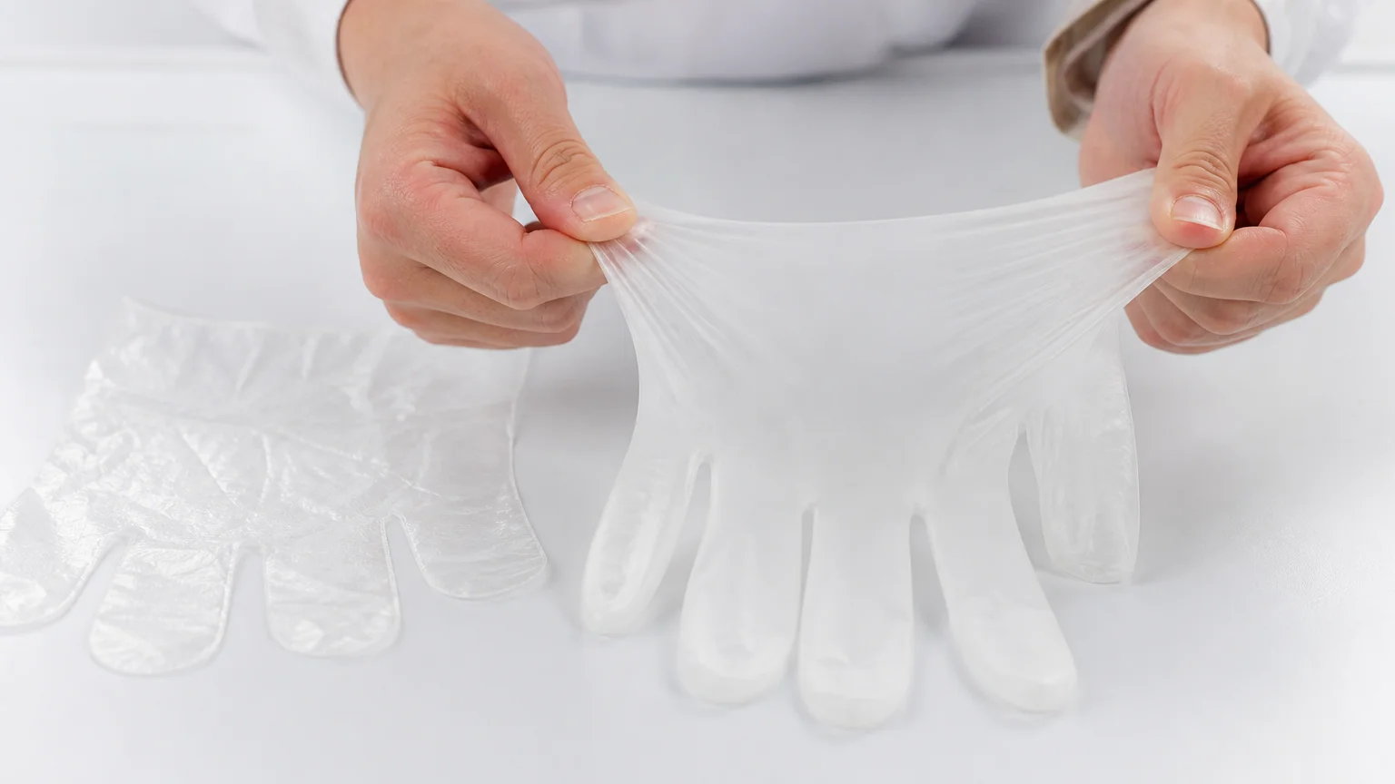 Close-up of a TPE glove being stretched to show elasticity beside a flat rigid PE glove on a white surface